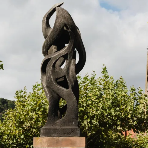 Eine moderne Skulptur aus schwarzem Material, die geschwungene Formen zeigt. Im Hintergrund sind Bäume und eine Kirche sichtbar.