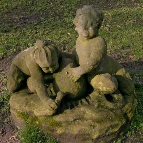 Eine steinerne Statue von zwei spielenden Kindern. Sie halten einen großen Ball und sind von grünem Gras umgeben.