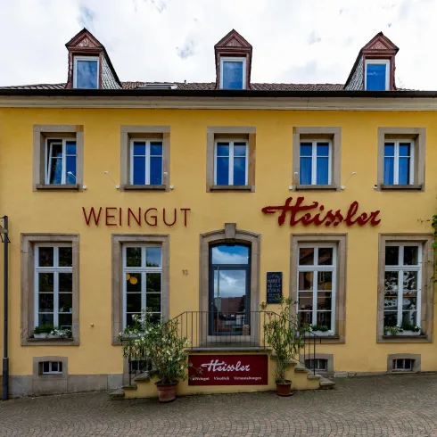Ein charmantes Weingut mit gelber Fassade und großen Fenstern. Die elegante Beschilderung "Heißler" ist gut sichtbar und von Pflanzen umgeben.