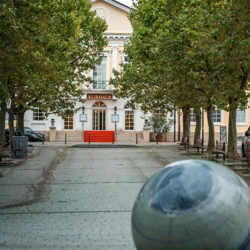 Eine ruhige Straße mit Bäumen führt zum Rathaus. Im Vordergrund befindet sich eine runde Skulptur.
