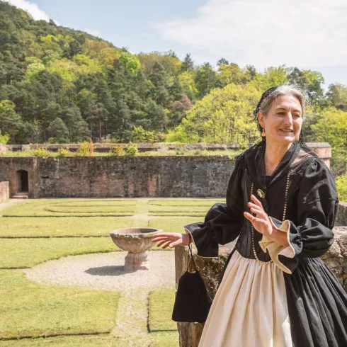 Eine Frau in historischer Kleidung steht in der Hardenburg bei Bad Dürkheim. Im Hintergrund sind grüne Wiesen und Bäume zu sehen.