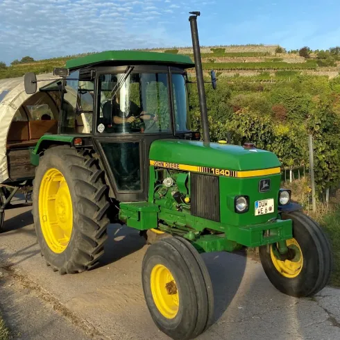 Ein grüner Traktor zieht einen Wagen mit Menschen durch die Weinberge. Die Umgebung ist grün und idyllisch mit sanften Hügeln im Hintergrund.