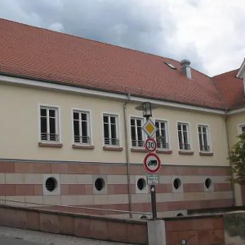 Ein modernes Gebäude mit einem roten Satteldach und großen Fenstern. Die Fassade kombiniert helle Farben mit kontrastierenden Elementen.