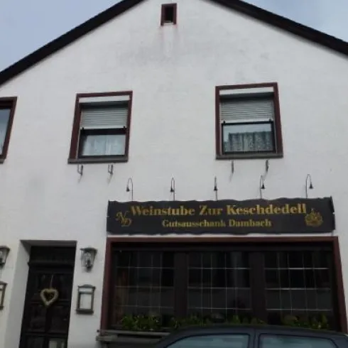 Ein charakteristisches Weinlokal mit der Beschriftung "Weinstube Zur Keschededell". Die Fassade ist schlicht und einladend, mit Fenstern und einer Eingangstür.