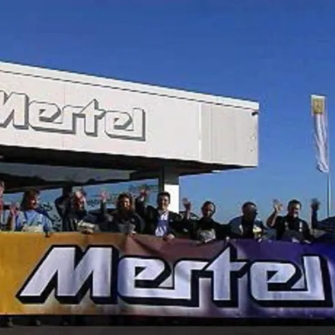 Eine Gruppe von Menschen steht vor einem Autohaus mit dem Namen "Mertel". Im Hintergrund sind die Logos von Renault und Dacia sowie einige Fahnen zu sehen.
