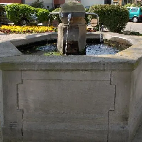 Ein schöner Steinbrunnen mit sprudelndem Wasser. Umgeben von gepflegten Grünflächen und bunten Blumen.