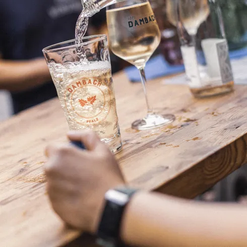 Ein Glas mit sprudelndem Getränk steht auf einer Holzbar. Im Hintergrund sind weitere Gläser und Menschen zu sehen.