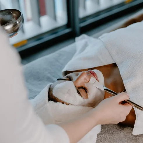 Eine Person genießt eine Gesichtsbehandlung in einem Spa. Die Therapeutin trägt eine Maske auf das Gesicht der liegenden Person auf.