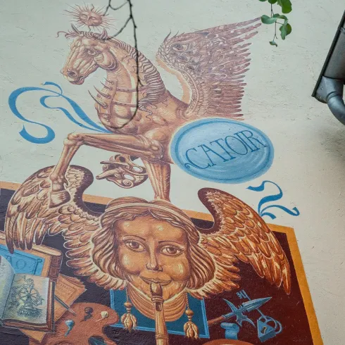 Ein farbenfrohes Wandgemälde zeigt ein geflügeltes Pferd und eine geflügelte Figur mit verschiedenen Symbolen. Das Kunstwerk ist an einer Hauswand zu sehen und ist von Pflanzen umgeben.