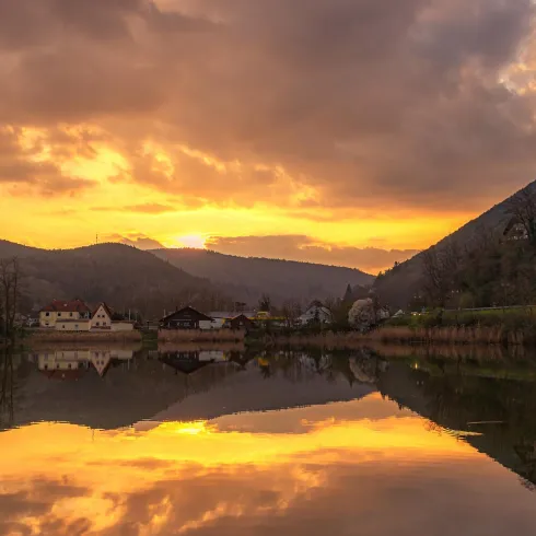 Ein ruhiger See spiegelt die beeindruckende Abenddämmerung wider. Sanfte Hügel und einige Häuser umrahmen die idyllische Landschaft.
