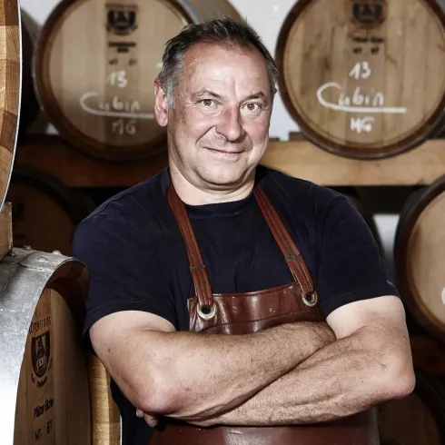 Ein Mann mit einem Lächeln steht vor Fässern aus Holz. Er trägt eine Schürze und strahlt Selbstbewusstsein aus.