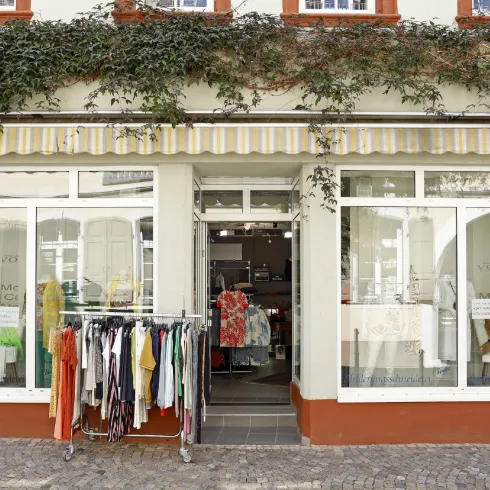 Ein Modegeschäft mit großen Fenstern und einer bunten Auswahl an Kleidung. Draußen stehen Kleider auf einem Ständer und die Schaufenster präsentieren weitere Modeartikel.