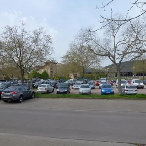 Ein großer Parkplatz mit vielen Autos. Im Hintergrund sind einige Bäume und ein Gebäude sichtbar.