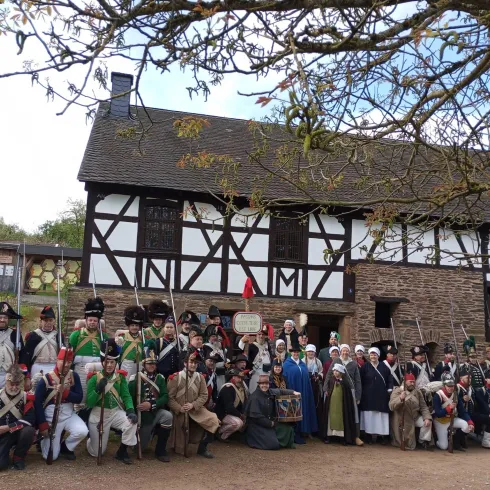 Eine Gruppe von Menschen in historischen Kostümen steht vor einem traditionellen Fachwerkhaus. Es scheint eine Nachbildung aus einer früheren Zeit zu sein, möglicherweise für eine Veranstaltung oder eine Reenactment-Darstellung.