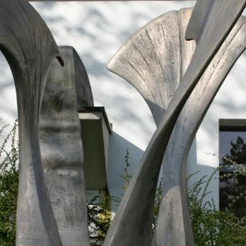 Eine moderne Skulptur aus Metall mit geschwungenen Formen steht vor einem Gebäude. Der Hintergrund zeigt eine Fensterscheibe und etwas grünes Pflanzenwachstum.