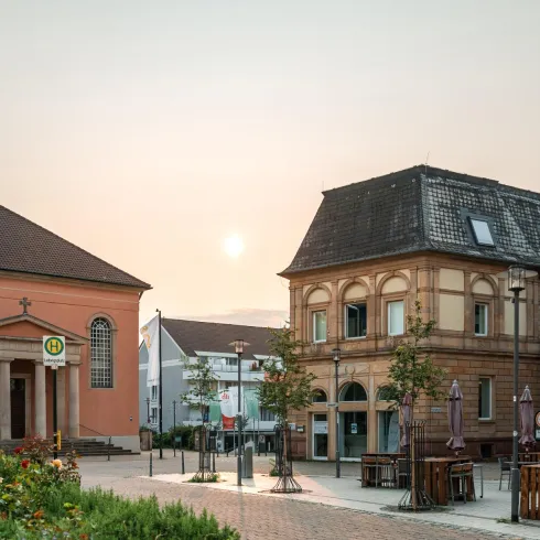 Ein malerischer Platz mit einer pinken Kirche und historischen Gebäuden. Die Sonne geht am Horizont auf und schafft eine ruhige Atmosphäre.