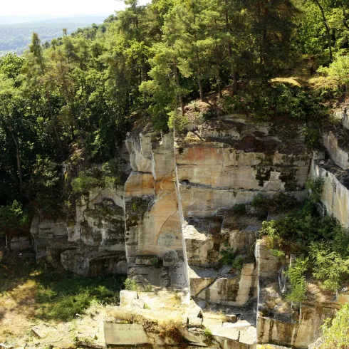 Eine beeindruckende Felslandschaft umgeben von Bäumen. Im Hintergrund ist eine Stadt sichtbar, die durch die Natur leicht versteckt wird.