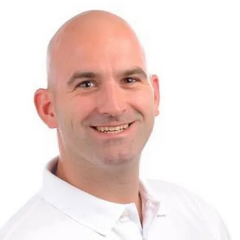 Ein Mann mit einer Glatze lächelt freundlich. Er trägt ein weißes Hemd und steht vor einem neutralen Hintergrund.