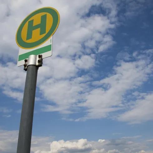 Ein Wartehäuschen mit einem gelben "H" für Haltestelle auf einem Metallpfosten. Der Himmel ist blau mit vielen Wolken.