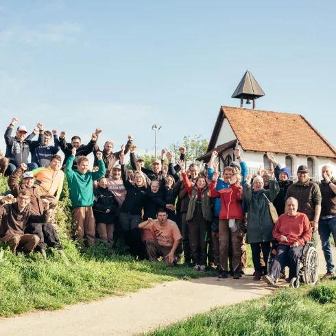Eine Gruppe von Menschen steht fröhlich vor einem kleinen Gebäude im Freien. Sie freuen sich und winken in die Kamera, umgeben von grünem Gras und blauem Himmel.