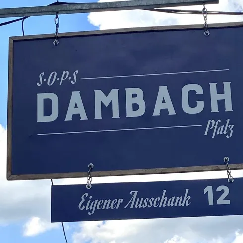 Ein Schild mit der Aufschrift "Dambach" und der Adresse "Eigener Ausschank 12". Der Himmel ist blau mit einigen Wolken.