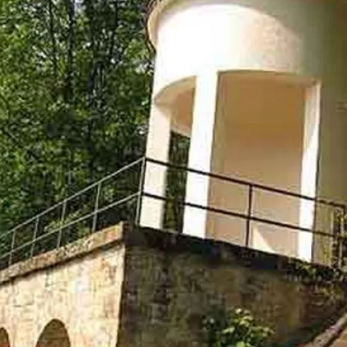 Ein historisches Bauwerk aus Stein mit einer weißen oberen Struktur steht in einer grünen Umgebung. Es hat eine Terrasse mit einem Geländer und ist von Bäumen umgeben.