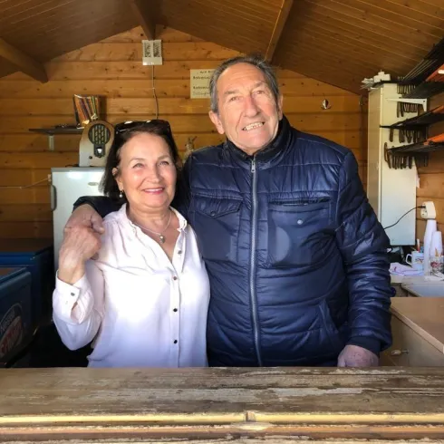 Ein fröhliches Paar steht hinter einem Holztresen in einem kleinen, gemütlichen Holzgebäude. Sie lächeln und posieren freundlich für das Foto.