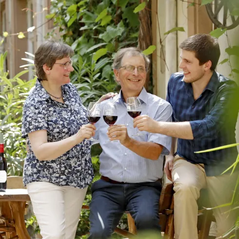 Eine fröhliche Gruppe von drei Personen hebt ihre Weingläser zum Anstoßen. Im Hintergrund sind Pflanzen und Weine auf einem Tisch zu sehen.