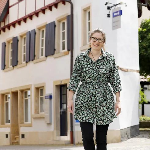 Eine Frau steht vor einem hübschen, traditionellen Gebäude mit Fensterläden. Sie trägt ein blumiges Kleid und lächelt freundlich.