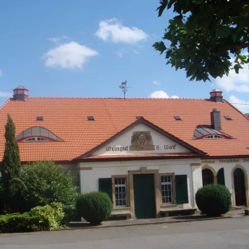 Ein Weingut mit einem schönen, roten Ziegeldach und einer einladenden Fassade. Umgeben von grünen Büschen und unter blauem Himmel.