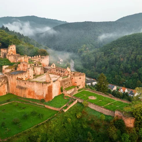 Eine alte Burruine umgeben von bewaldeten Hügeln und sanften Nebelschwaden. Der grüne Rasen und die teilweise erhaltenen Mauern verleihen der Szenerie noch mehr Charme.