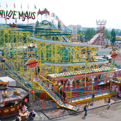 Eine bunte Achterbahn namens "Wilde Maus" in einem Freizeitpark. Viele Menschen genießen die Attraktionen und die fröhliche Atmosphäre.