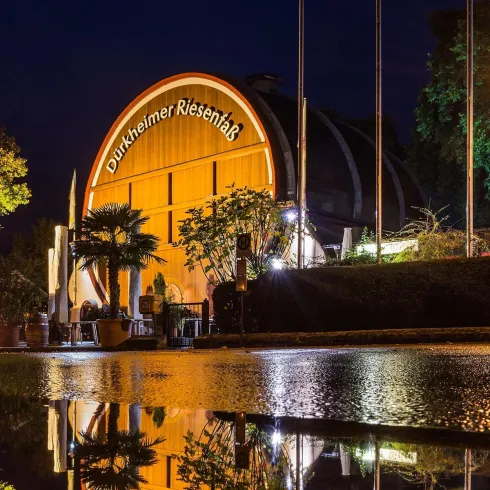 Ein großes, rundes Gebäude mit der Aufschrift "Dürheimer Riesenrad" ist zwischen Bäumen zu sehen. Im Hintergrund ist ein Uhrturm und weitere Gebäude zu erkennen.
