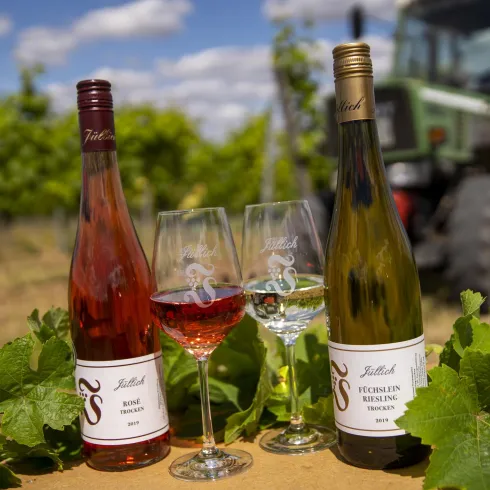 Zwei Flaschen Wein stehen neben Weingläsern auf einem Tisch. Im Hintergrund sind Weinreben und ein Traktor zu sehen.