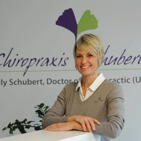 Eine freundliche Chiropraktikerin steht an einem Empfangstresen. Im Hintergrund ist das Logo der Chiropraktikpraxis zu sehen.