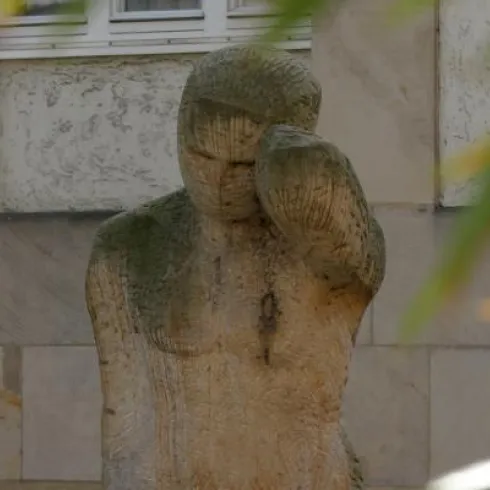 Eine statue eines nachdenklichen Menschen aus Stein. Die Figur hat eine ruhige Ausstrahlung und steht im Freien.