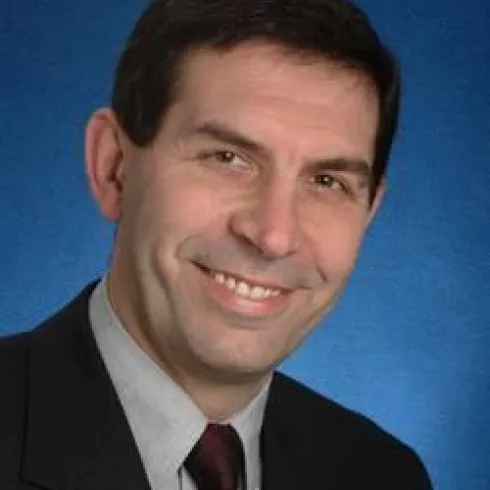 Ein lächelnder Mann in einem Anzug vor einem blauen Hintergrund. Er hat kurze, dunkle Haare und trägt eine Krawatte.