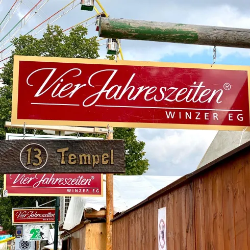 Ein auffälliges Schild der „Vier Jahreszeiten Winzer EG“ mit dem Hinweis „Tempel“. Im Hintergrund ist ein Riesenrad und eine rustikale Holzkonstruktion zu sehen.
