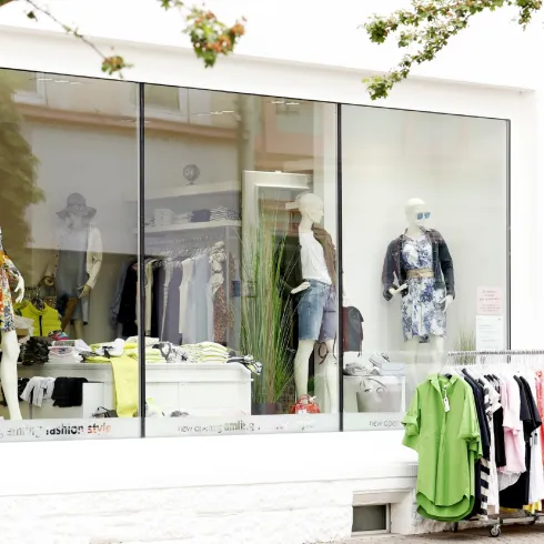 Ein Schaufenster mit Modellen, die verschiedene Kleidungsstücke präsentieren. Darunter befindet sich ein Kleiderständer mit farbenfrohen Outfits.