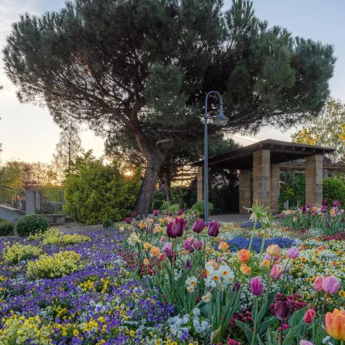 Ein schöner Garten mit bunten Blumen und einem kleinen Holzbrücke. Im Hintergrund sind Bäume und ein überdachter Sitzbereich sichtbar.