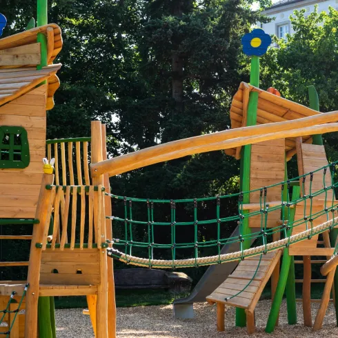 Ein Spielplatz mit Holzkonstruktionen, einer Brücke und bunten Blumen. Im Hintergrund sind Bäume und ein Gebäude zu sehen.