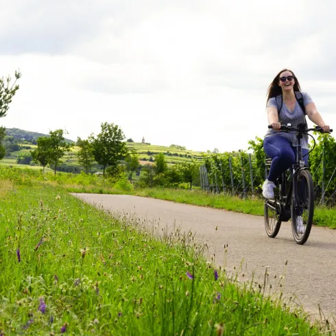 Eine Frau fährt fröhlich mit dem Fahrrad auf einem Weg durch Weinberge. Die Landschaft ist grün und bietet einen schönen Ausblick auf Hügel und Bäume.