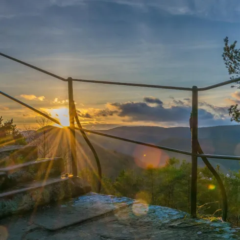 Ein schöner Blick von einem Felsen mit Sonnenstrahlen, die durch die Bäume scheinen. Im Hintergrund sind sanfte Hügel und ein klarer Himmel zu sehen.