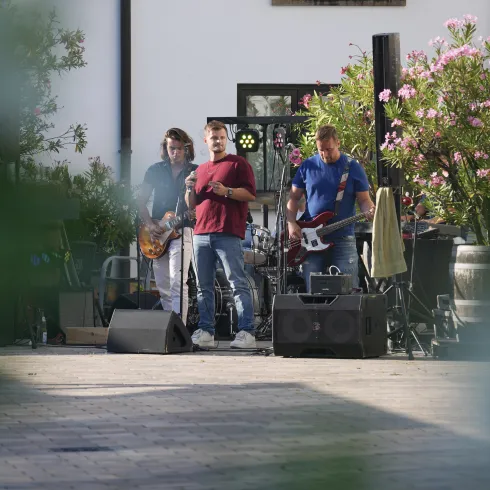 Eine Band spielt live auf einer Freiluftbühne. Im Hintergrund sind grüne Pflanzen und Blumen zu sehen.