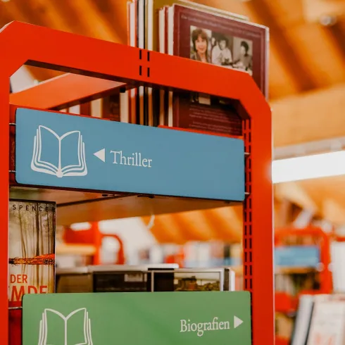 Ein Bücherregal in einer Bibliothek mit den Beschilderungen für die Kategorien "Thriller" und "Biografien". Die Regale sind rot und die Umgebung wirkt freundlich und einladend.