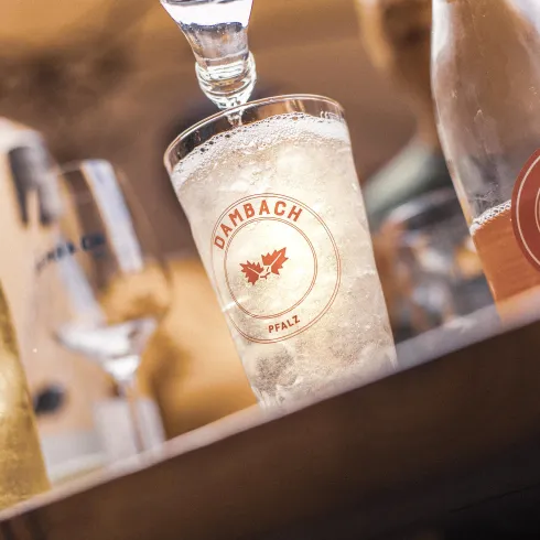 Ein Glas mit sprudelndem Getränk steht auf einem Tisch, umgeben von Flaschen. Der Fokus liegt auf dem Glas mit dem Dambach-Logo.