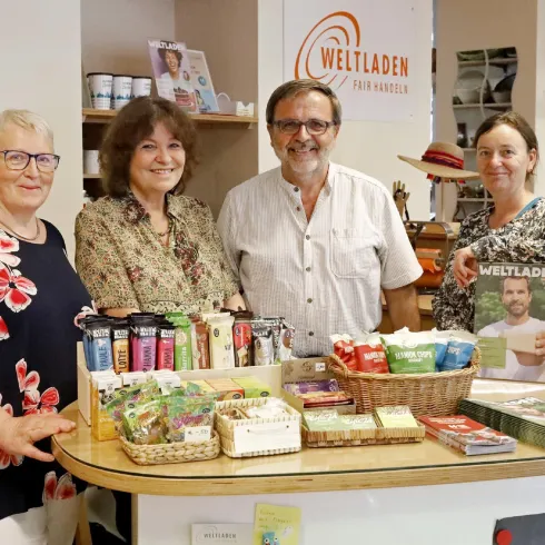 Ein freundliches Team steht hinter einem Verkaufstresen im Weltladen. Auf dem Tisch sind verschiedene Produkte wie Getränke und Snacks aus fairem Handel ausgestellt.