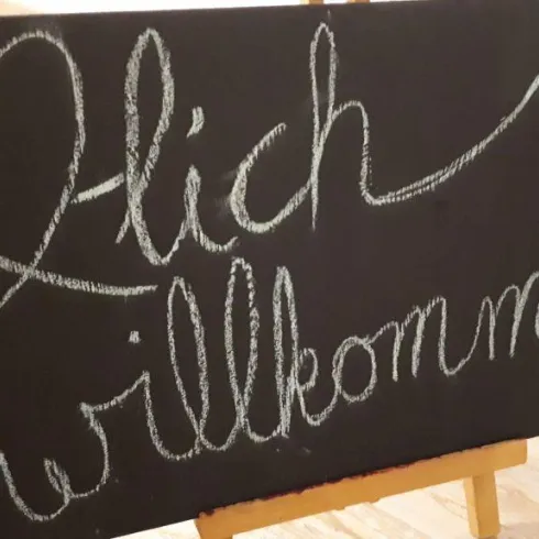 Ein schwarzes Schild mit der Aufschrift "Herzlich willkommen" in freundlicher Schrift. Daneben ist eine einfache Zeichnung einer Sonne zu sehen.