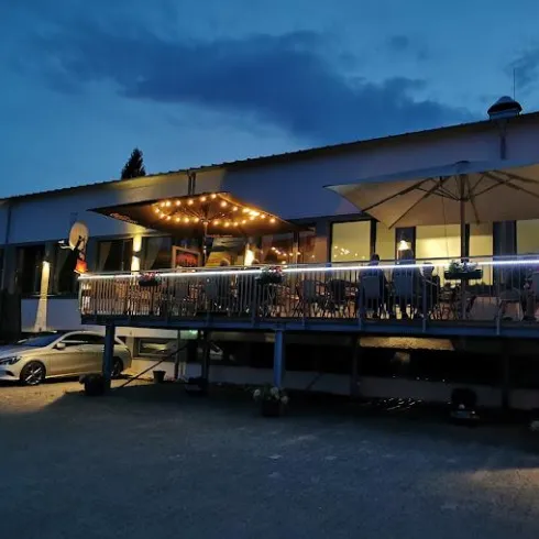 Ein modernes Restaurant mit einer beleuchteten Terrasse. Im Vordergrund steht ein Auto und die Umgebung ist abends ruhig.
