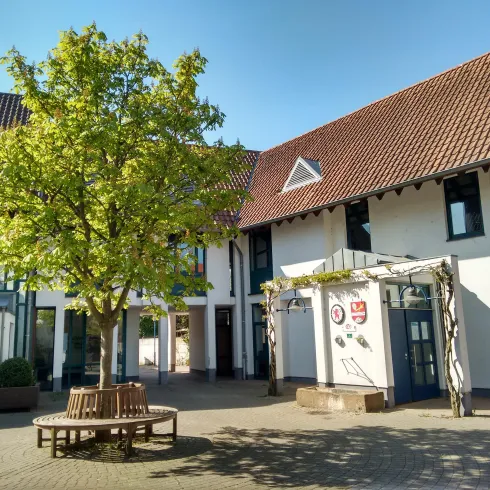 Ein schöner Innenhof mit einem großen Baum und modernen Gebäuden. Die Sonne scheint und es ist ein klarer blauer Himmel zu sehen.